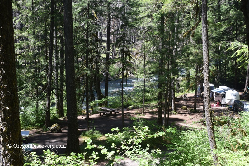 Olallie Campground in Oregon: The picture "Olallie Campground riverside campsites" shows a shaded forested area with picnic tables and several campers or trailers set up among tall trees. The clear river can be seen running behind the campsites.