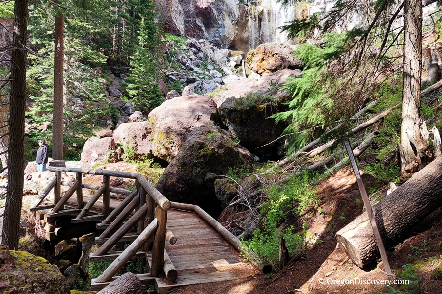 Paulina Falls in Oregon - Paulina Falls Wooden Bridge Paulina Falls in Oregon: The picture "Paulina Falls Wooden Bridge" shows a rustic wooden footbridge leading over a small creek toward massive boulders at the base of the falls, with a person admiring the view in the distance.
