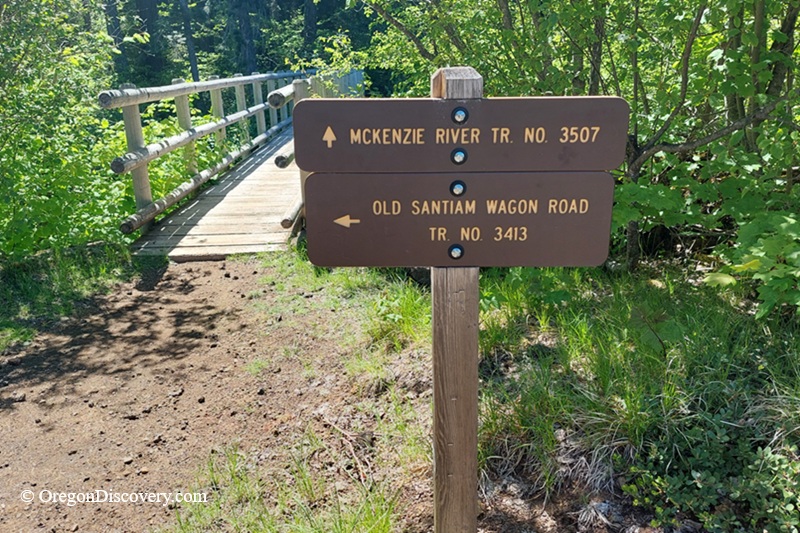 McKenzie River Trail in Oregon: The picture "McKenzie River Trail and Old Santiam Wagon Road junction sign" displays a brown wooden sign marking the start of McKenzie River Trail No. 3507 and Old Santiam Wagon Road Trail No. 3413, standing at the edge of a wooden bridge in a lush forest setting.
