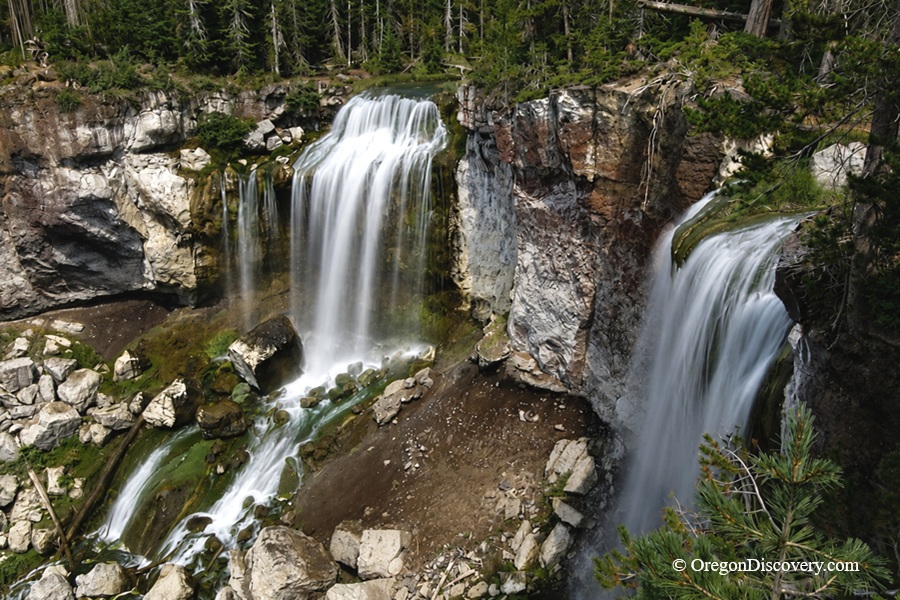 Paulina Falls in Oregon - Paulina Falls Aerial Paulina Falls in Oregon: The picture "Paulina Falls Aerial" features a dramatic high-angle view of two powerful waterfalls side by side, plunging into a rocky canyon surrounded by evergreen trees and multicolored cliffs.