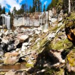 Paulina Falls in Oregon - Paulina Falls Base Paulina Falls in Oregon: The picture "Paulina Falls Base" presents a wide view of the waterfall base, with white water streaming down a tall cliff face amid a landscape of large rocks, logs, and green foliage.
