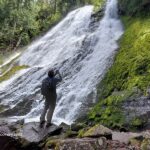 Diamond Creek Falls in Oregon: The picture "Diamond Creek Falls Close-up" shows a person standing on rocks at the base of the tall, sloped waterfall, taking a photo of the water streaming over mossy rocks and the surrounding vibrant greenery.
