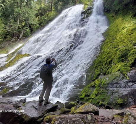 Diamond Creek Falls in Oregon: The picture "Diamond Creek Falls Close-up" shows a person standing on rocks at the base of the tall, sloped waterfall, taking a photo of the water streaming over mossy rocks and the surrounding vibrant greenery.