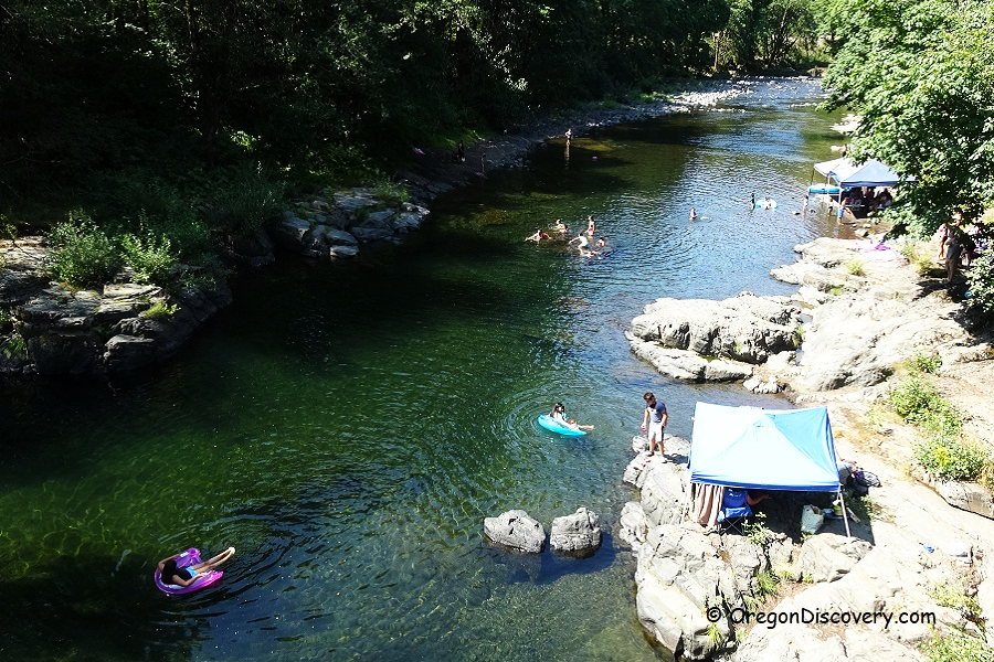 Jones Creek Day-Use Area and Campground in Oregon: The picture "Jones Creek Swimming Hole" shows people swimming and floating on inner tubes in a clear, green river surrounded by forest, with rocky banks and a blue canopy tent set up along the shore.