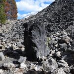 Big Obsidian Flow in Oregon: The picture "Obsidian Monolith" shows an imposing, upright black obsidian boulder standing among a scatter of volcanic rocks at the base of a rocky hill, with trees in the background.