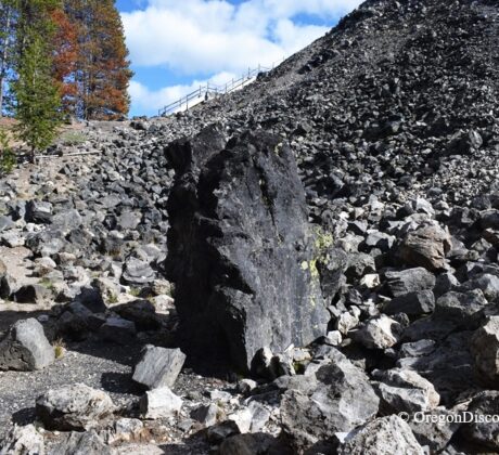 Big Obsidian Flow in Oregon: The picture "Obsidian Monolith" shows an imposing, upright black obsidian boulder standing among a scatter of volcanic rocks at the base of a rocky hill, with trees in the background.