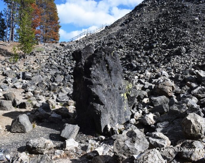 Big Obsidian Flow in Oregon: The picture "Obsidian Monolith" shows an imposing, upright black obsidian boulder standing among a scatter of volcanic rocks at the base of a rocky hill, with trees in the background.