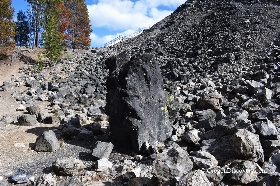 Big Obsidian Flow in Oregon: The picture "Obsidian Monolith" shows an imposing, upright black obsidian boulder standing among a scatter of volcanic rocks at the base of a rocky hill, with trees in the background.