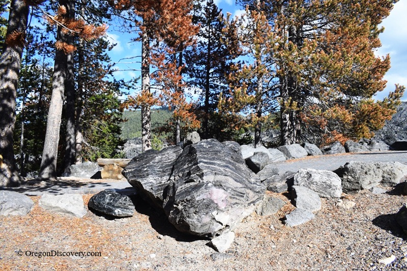 Big Obsidian Flow in Oregon: The picture "Obsidian Boulder" displays a large, shiny black obsidian rock surrounded by smaller stones and pine trees, with dappled sunlight on the ground.