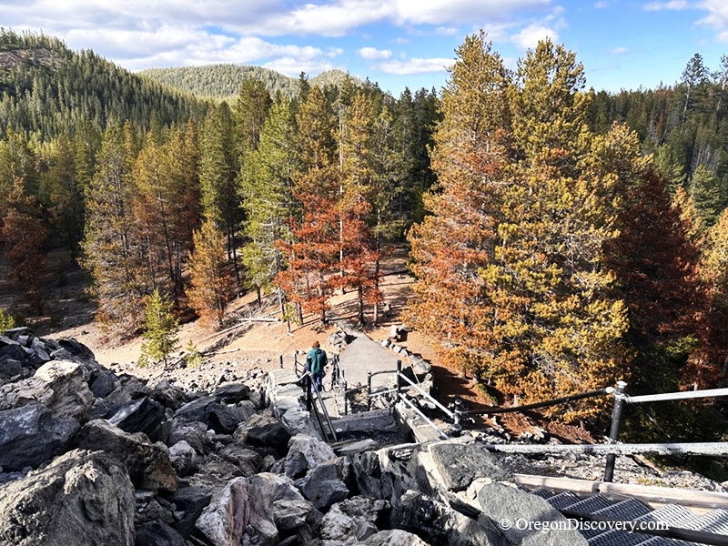 Big Obsidian Flow in Oregon: The picture "Obsidian Flow Forest Edge" shows a hiker walking down stairs from the rocky flow into a forest of green and autumn-colored pine trees, with mountains in the distance.