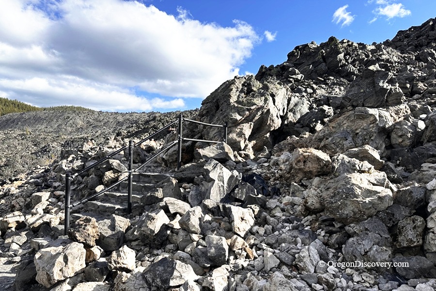 Big Obsidian Flow in Oregon: The picture "Obsidian Trail Staircase" features a rocky staircase with black railings climbing up a slope of large obsidian and pumice rocks beneath a vivid blue sky.