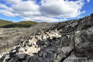 Big Obsidian Flow in Oregon: The picture "Big Obsidian Flow Trail" shows a rugged hiking path winding through a vast field of jagged volcanic rocks with mountains and a partly cloudy blue sky in the background.
