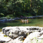 Jones Creek Day-Use Area and Campground in Oregon: The picture "Jones Creek River Rocks" features large gray rocks in the foreground with families wading and swimming in the shallow river, bordered by lush green trees along the banks.