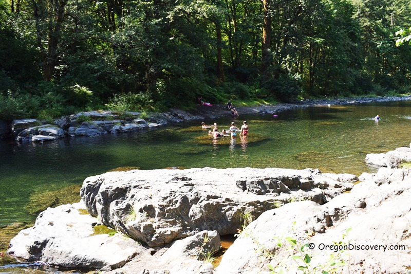 Jones Creek Day-Use Area and Campground in Oregon: The picture "Jones Creek River Rocks" features large gray rocks in the foreground with families wading and swimming in the shallow river, bordered by lush green trees along the banks.