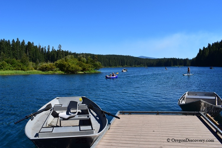 Clear Lake in Oregon – Clear Lake Dock and Boaters Clear Lake in Oregon – The picture "Clear Lake Dock and Boaters" shows a rowboat tied to a dock in the foreground with people in paddle boats and kayaks enjoying the clear blue lake, all surrounded by lush green forest and mountains under a bright blue sky.