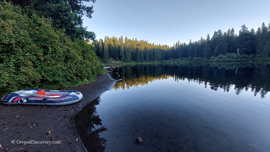 Clear Lake in Oregon – Clear Lake Beach at Dusk Clear Lake in Oregon – The picture "Clear Lake Beach at Dusk" shows a quiet, tree-lined shoreline with a small inflatable raft on the dark sand, and the tranquil lake surface reflecting fir trees in the soft evening light.