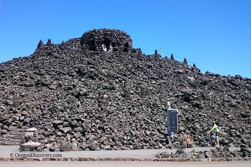 Dee Wright Observatory in Oregon – Dee Wright Observatory Lava MoundDee Wright Observatory in Oregon – The picture "Dee Wright Observatory Lava Mound" displays the observatory as a rugged stone structure built on top of a mound of dark lava rocks, with people and cyclists standing near the base and at the entrance under a clear blue sky.