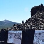 Dee Wright Observatory in Oregon – Dee Wright Observatory Interpretation Signs Dee Wright Observatory in Oregon – The picture "Dee Wright Observatory Interpretation Signs" shows scenic outdoor interpretive signs about local history and geology in front of jagged lava rock formations, with forested mountains and a bright sky beyond.