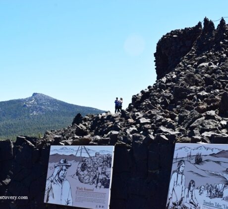 Dee Wright Observatory in Oregon – Dee Wright Observatory Interpretation Signs Dee Wright Observatory in Oregon – The picture "Dee Wright Observatory Interpretation Signs" shows scenic outdoor interpretive signs about local history and geology in front of jagged lava rock formations, with forested mountains and a bright sky beyond.