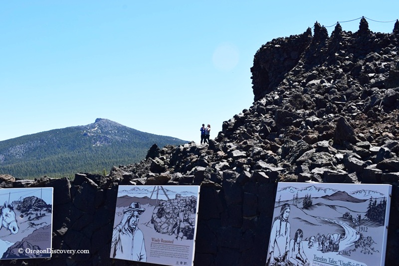 Dee Wright Observatory in Oregon – Dee Wright Observatory Interpretation SignsDee Wright Observatory in Oregon – The picture "Dee Wright Observatory Interpretation Signs" shows scenic outdoor interpretive signs about local history and geology in front of jagged lava rock formations, with forested mountains and a bright sky beyond.