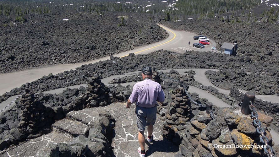 Dee Wright Observatory in Oregon – Dee Wright Observatory Viewpoint Path Dee Wright Observatory in Oregon – The picture "Dee Wright Observatory Viewpoint Path" captures a person walking down a stone and gravel path surrounded by rugged black lava rocks, with a parking lot, cars, and dense forest in the background.