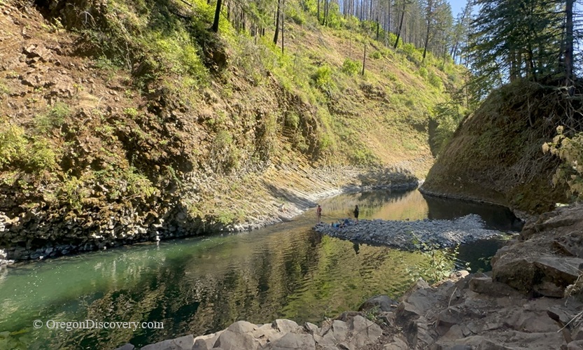 Molalla Eye, Oregon: Scenic Swimming & Fishing Spot on the Molalla River – Molalla Eye Swimming Hole Molalla Eye, Oregon: Scenic Swimming & Fishing Spot on the Molalla River – The picture "Molalla Eye Swimming Hole" shows a peaceful bend in the river, with people standing on a rocky islet in the emerald-green water, surrounded by steep basalt cliffs and sunlit fir trees.
