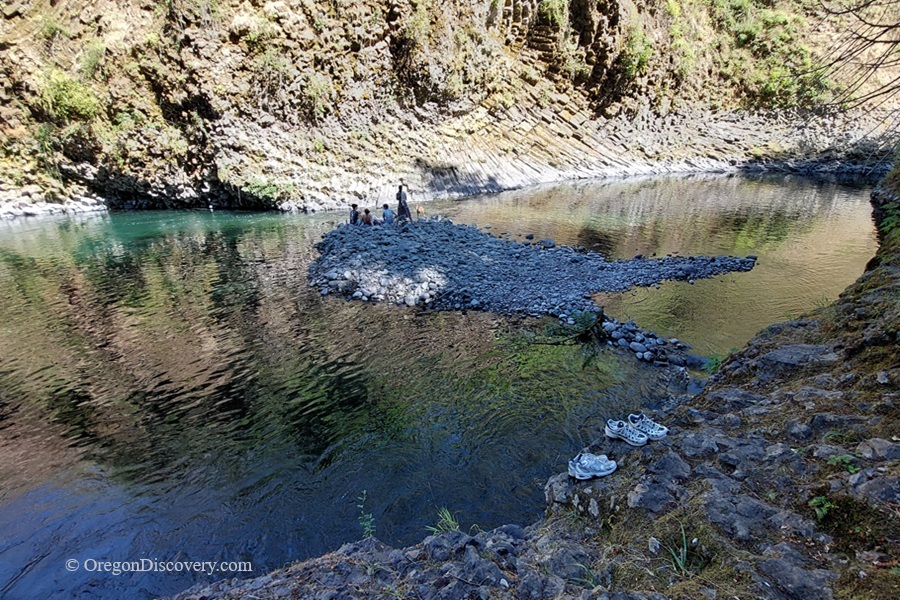 Molalla Eye, Oregon: Scenic Swimming & Fishing Spot on the Molalla River – Molalla Eye Pebble BarMolalla Eye, Oregon: Scenic Swimming & Fishing Spot on the Molalla River – The picture "Molalla Eye Pebble Bar" captures a small group of people gathered on a gray pebble bar in the middle of the gently flowing river, with layered rock walls rising steeply and shoes sitting on the rocks in the foreground