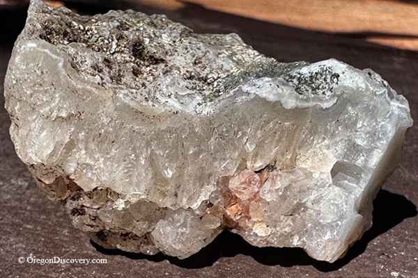 Rooster Rock Rockhounding in Oregon – Rooster Rock Agate Crystal Close-upRooster Rock Rockhounding in Oregon – The picture "Rooster Rock Agate Crystal Close-up" shows a detailed view of a chunky agate and quartz crystal specimen with layered, translucent bands and mineral inclusions, resting on a sunlit surface.