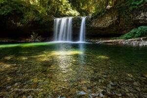 Upper Butte Creek Falls Oregon – Upper Butte Creek Falls Long Exposure Upper Butte Creek Falls Oregon: The picture "Upper Butte Creek Falls Long Exposure" shows a long-exposure view of the waterfall, giving the falling water a silky effect as it pours into a vibrant green forest pool with a stony bottom and logs at the water’s edge.