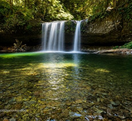 Upper Butte Creek Falls Oregon – Upper Butte Creek Falls Long Exposure Upper Butte Creek Falls Oregon: The picture "Upper Butte Creek Falls Long Exposure" shows a long-exposure view of the waterfall, giving the falling water a silky effect as it pours into a vibrant green forest pool with a stony bottom and logs at the water’s edge.