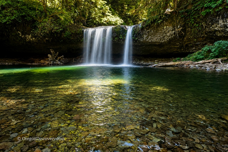 Upper Butte Creek Falls Oregon – Upper Butte Creek Falls Long Exposure Upper Butte Creek Falls Oregon: The picture "Upper Butte Creek Falls Long Exposure" shows a long-exposure view of the waterfall, giving the falling water a silky effect as it pours into a vibrant green forest pool with a stony bottom and logs at the water’s edge.