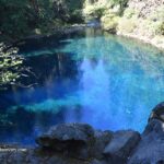 Tamolich Blue Pool (Oregon's Blue Pool) in Oregon – Blue Pool Swimmers and Rocky Shore Tamolich Blue Pool (Oregon's Blue Pool) in Oregon – The picture "Blue Pool Swimmers and Rocky Shore" features people sitting and relaxing on rocks beside the deep, clear blue water of the pool, with rocky cliffs and reflections of trees in the background.