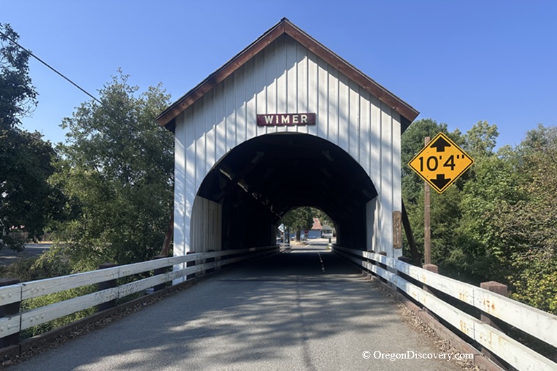 Wimer Covered Bridge in Oregon – Wimer Covered Bridge EntranceWimer Covered Bridge in Oregon – The picture "Wimer Covered Bridge Entrance" shows the front view of the white wooden Wimer Covered Bridge with a peaked roof, an entrance sign, and a yellow clearance sign reading "10'4"". Trees frame the bridge on either side.