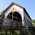 Wimer Covered Bridge in Oregon – Wimer Covered Bridge Side View Wimer Covered Bridge in Oregon – The picture "Wimer Covered Bridge Side View" features a side perspective of the historic wooden bridge, raised on concrete supports above a green riverbank, with diagonal brown and white guard rails and a yellow clearance sign on the entrance.