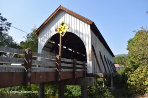 Wimer Covered Bridge in Oregon – Wimer Covered Bridge Side View Wimer Covered Bridge in Oregon – The picture "Wimer Covered Bridge Side View" features a side perspective of the historic wooden bridge, raised on concrete supports above a green riverbank, with diagonal brown and white guard rails and a yellow clearance sign on the entrance.