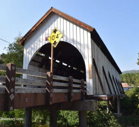 Wimer Covered Bridge in Oregon – Wimer Covered Bridge Side View Wimer Covered Bridge in Oregon – The picture "Wimer Covered Bridge Side View" features a side perspective of the historic wooden bridge, raised on concrete supports above a green riverbank, with diagonal brown and white guard rails and a yellow clearance sign on the entrance.
